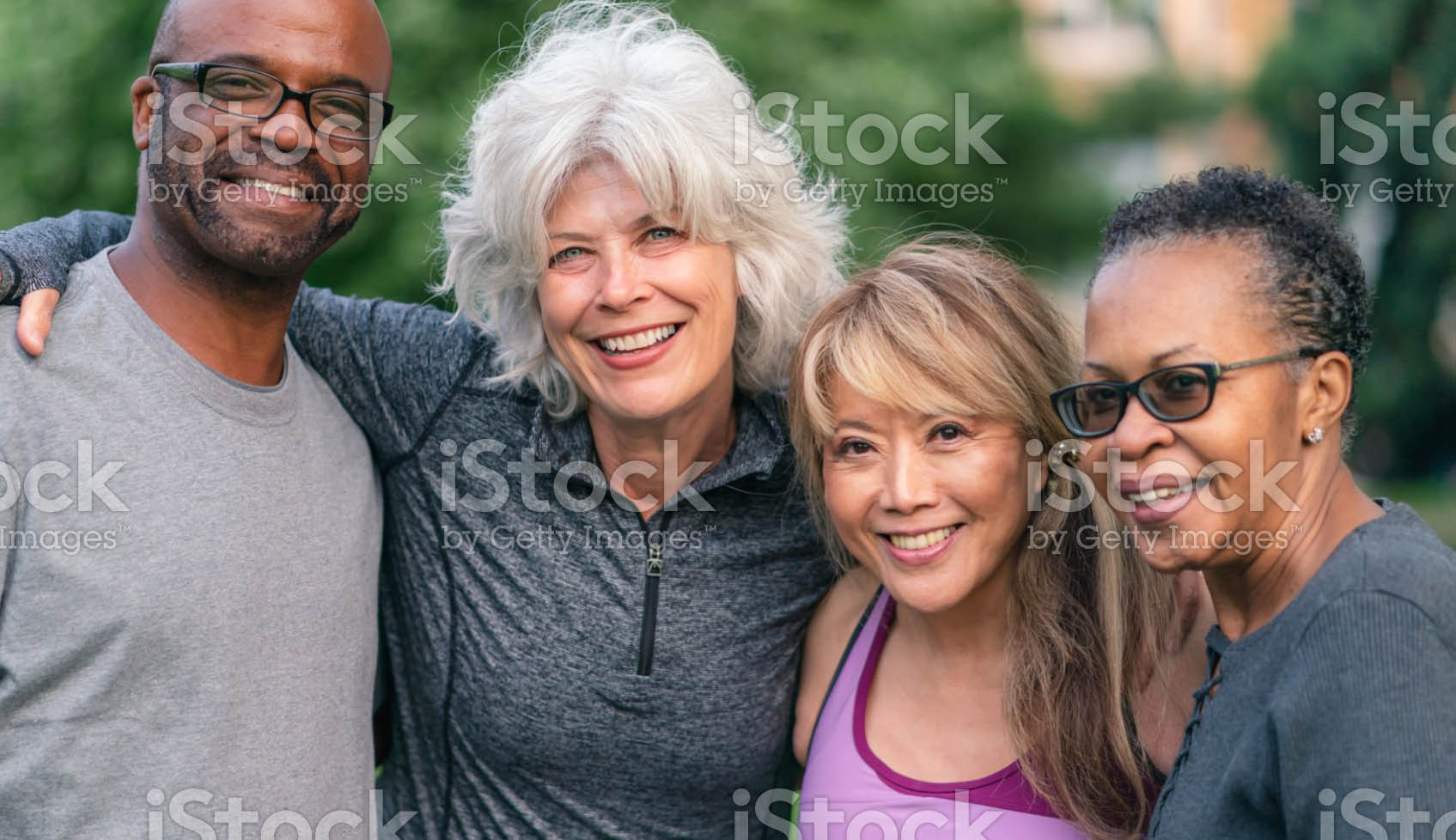 Portrait of a multi-ethnic group of senior friends at a park. The individuals are standing with arms around each others shoulders. They are smiling confidently directly at the camera. They are wearing casual athletic clothing.