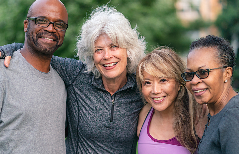 Portrait of a multi-ethnic group of senior friends at a park. The individuals are standing with arms around each others shoulders. They are smiling confidently directly at the camera. They are wearing casual athletic clothing.