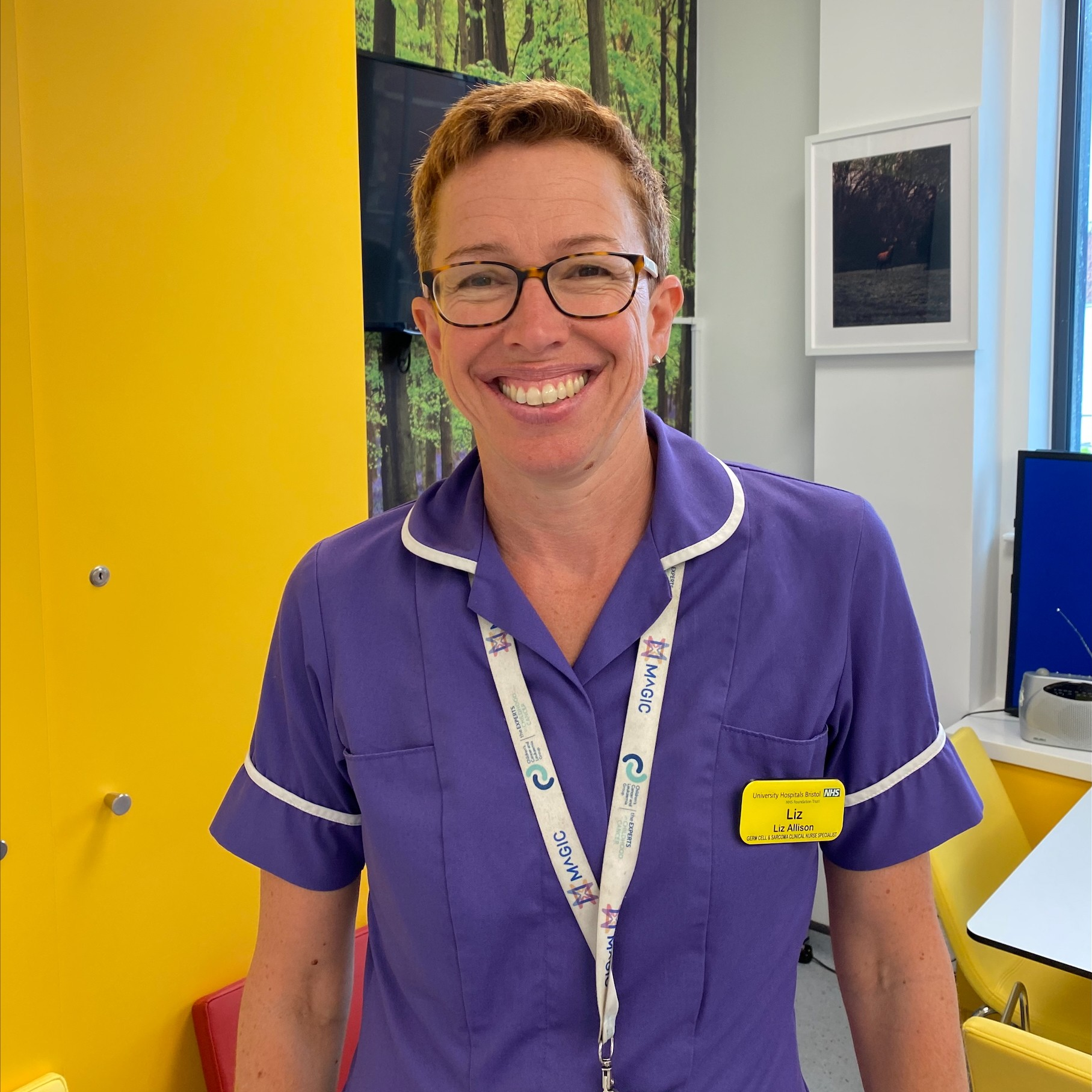 This photo is of Lead Sarcoma Clinical Nurse Specialist Liz Allison. A woman wearing an NHS uniform with white trim, a lanyard, and an ID badge stands in a room with yellow and green walls.